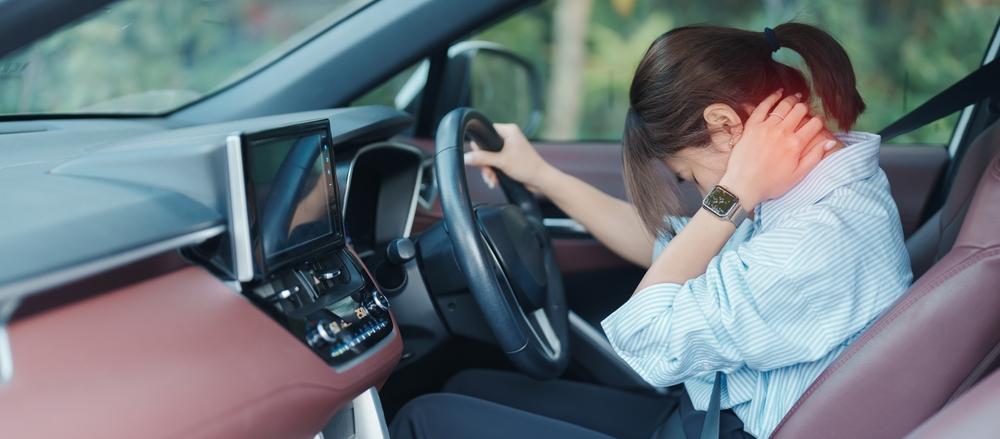 A woman sits in a car's driver's seat, resting her head on the steering wheel, appearing tired or contemplative.