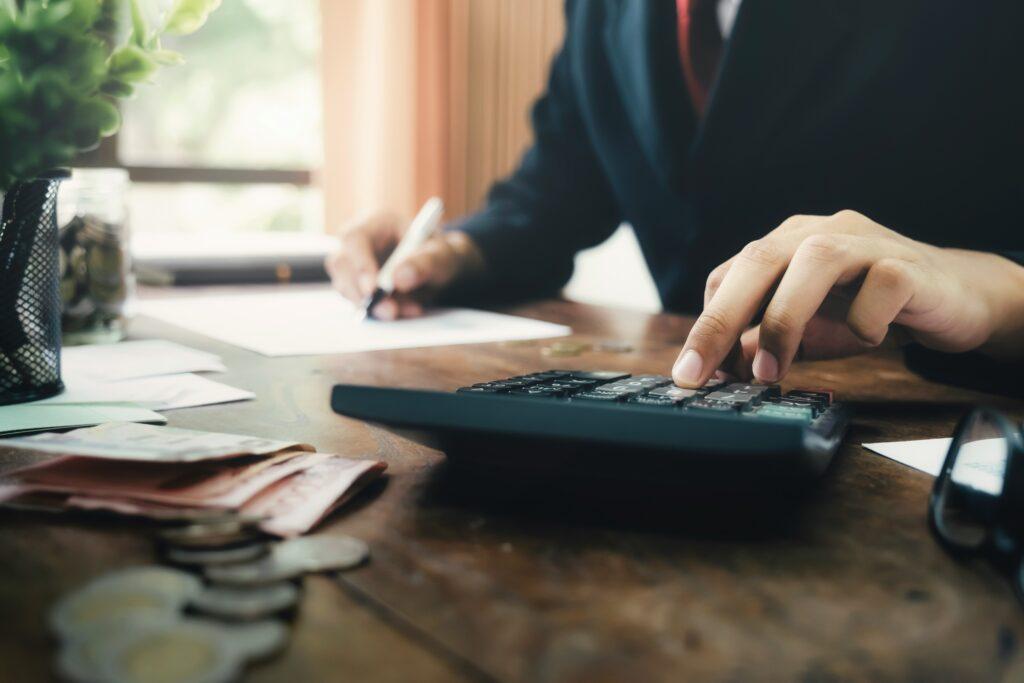 A person in business attire sits at a wooden desk, using a calculator with one hand while writing on paperwork with the other. Banknotes and coins are scattered in the foreground, giving the scene a focused, financial-work atmosphere bathed in soft natural light.