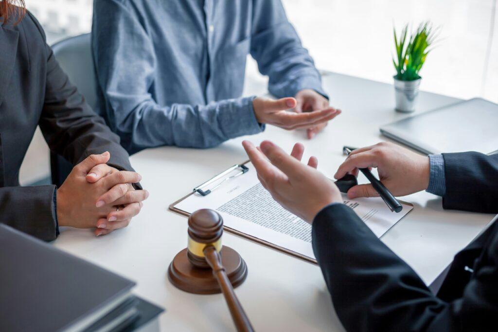A group sits around a bright office table, their hands mid-gesture as they discuss paperwork clipped to a clipboard. A wooden gavel rests nearby, emphasizing the legal setting, while a small plant and laptop sit in the background.