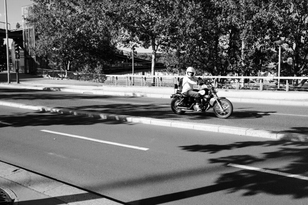 A motorcyclist wearing a helmet rides along a sunlit road, casting strong shadows across the pavement. Trees line the background, and the bright daylight creates sharp contrasts in this black-and-white scene.