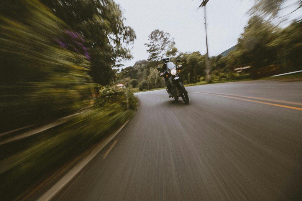 A motorcyclist rides quickly along a curving road surrounded by trees, with the background stretched into dramatic motion blur that emphasizes speed and the sweeping path ahead.