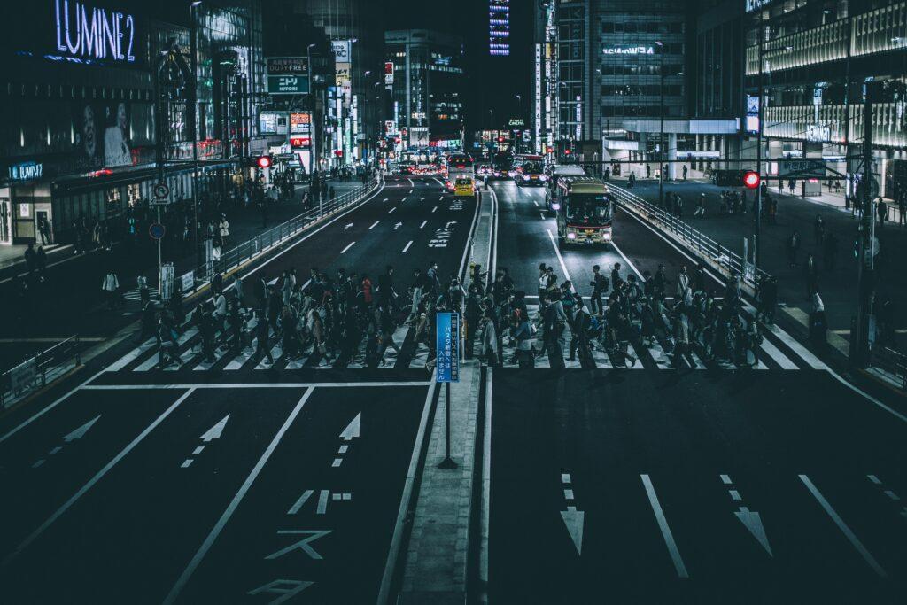 A crowded nighttime crosswalk in a brightly lit city, with large groups of pedestrians crossing multiple lanes as buses and cars wait under glowing signs and neon lights.