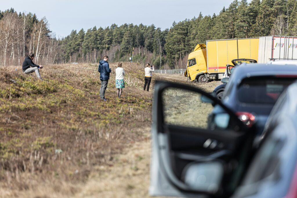 A roadside crash scene with a yellow cargo truck stuck in a grassy embankment, several people standing nearby observing or taking photos, and a parked car with its door open in the foreground, all set against a backdrop of sparse trees and a clear sky.