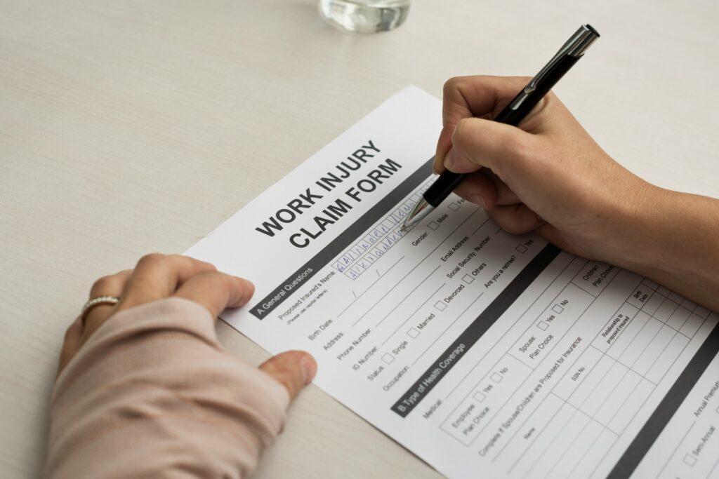 A person filling out a work injury claim form at a desk, holding a pen and writing personal details on the document.