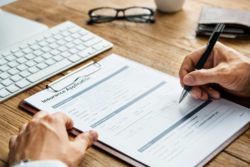 A close-up of someone completing an insurance application form on a clipboard at a wooden desk, with a keyboard, glasses, and a wallet nearby.