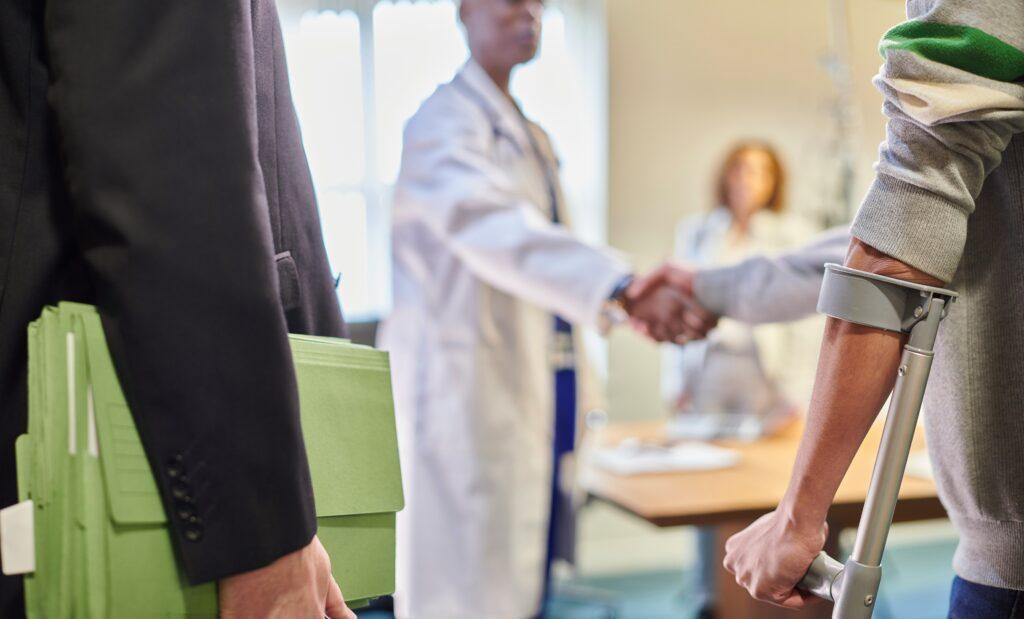An injured person on crutches shakes hands with a doctor in a bright office, while someone in a suit holds green case files in the foreground, suggesting a medical or legal consultation.