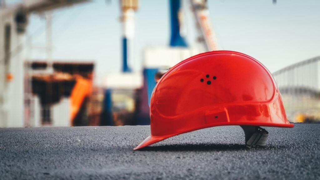 A bright red construction hard hat lying on the ground at an outdoor worksite with blurred industrial equipment in the background.