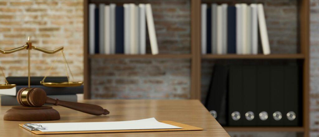 Wooden judge’s gavel resting on a desk beside a blank document on a clipboard, with a brass balance scale in the background and shelves filled with neatly arranged books and black binders against an exposed brick wall, creating a formal legal office or courtroom setting with warm lighting and a shallow depth of field.