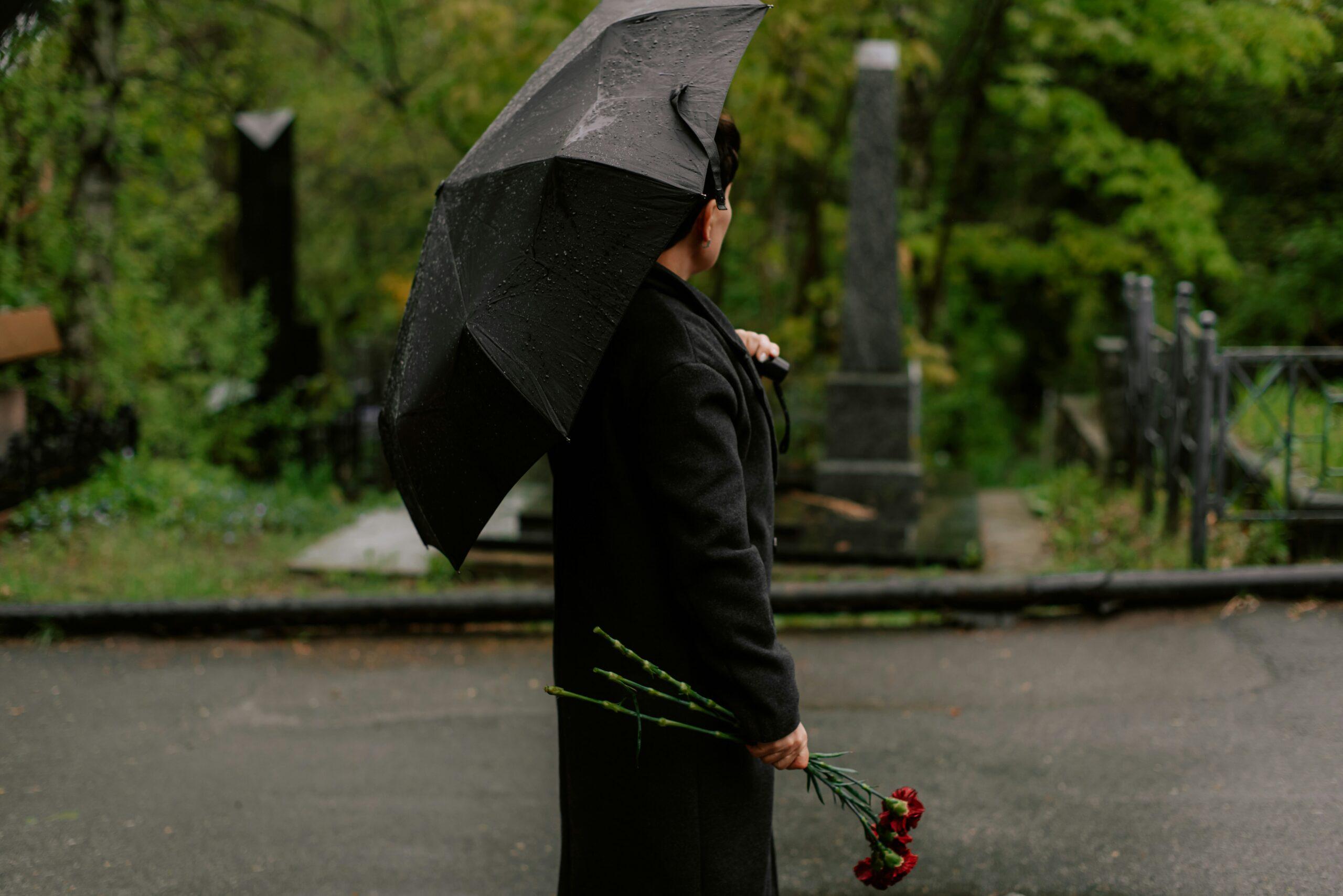 Person dressed in dark clothing standing on a rain-wet path in a cemetery, holding a black umbrella angled over their shoulder and a small bouquet of red carnations hanging downward in one hand, with gravestones, stone monuments, and iron fencing blurred in the background, green trees and foliage surrounding the scene, and an overcast, somber atmosphere suggesting mourning or remembrance.